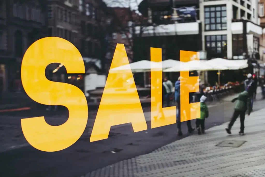 Gold and black sale sign in store window reflecting people walking by on sidewalk outside